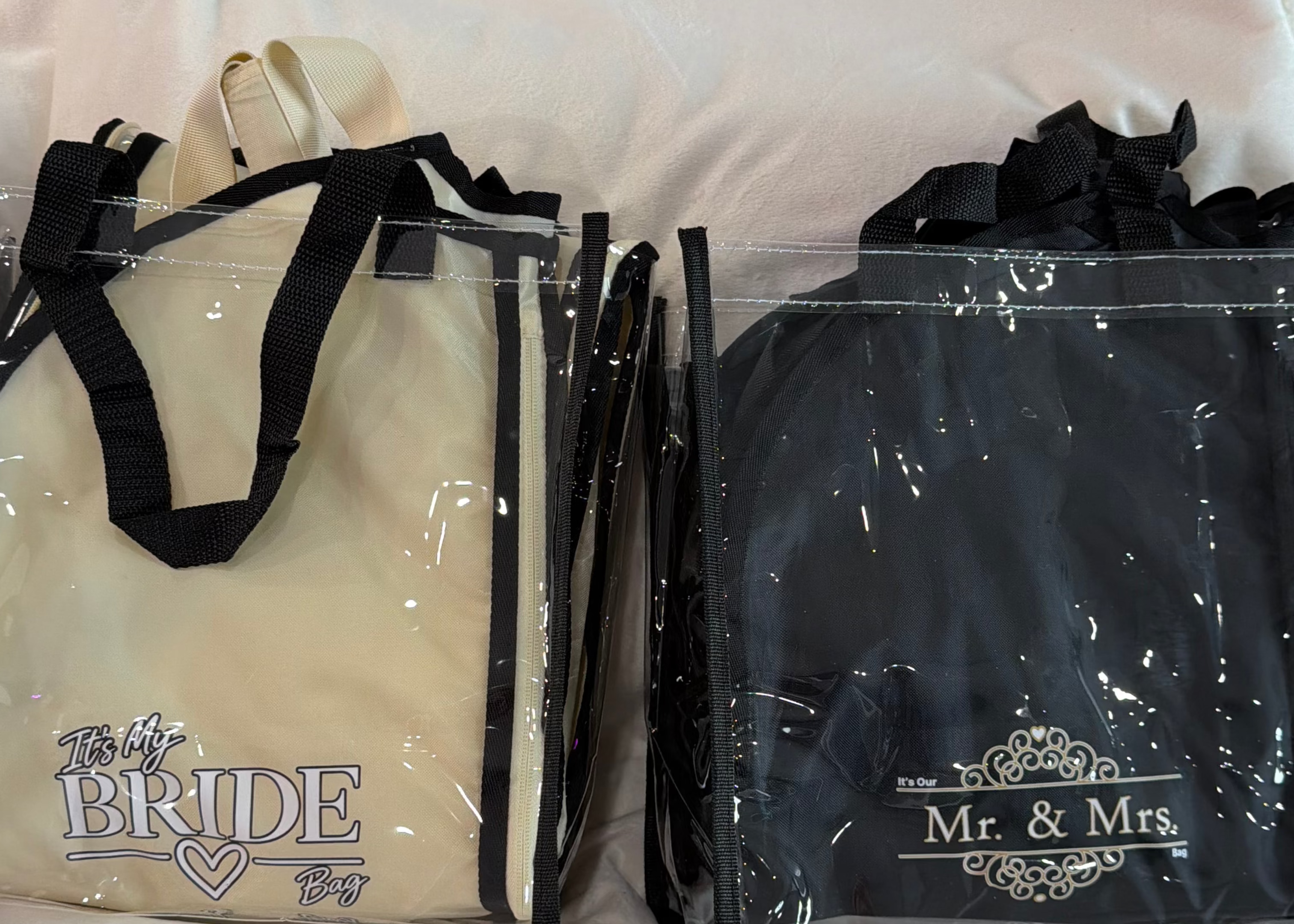 Two clear plastic tote bags with black handles and trim, placed on a white blanket. The left bag is cream with 'The Bride' printed on it, and the right bag is black with 'Mr.